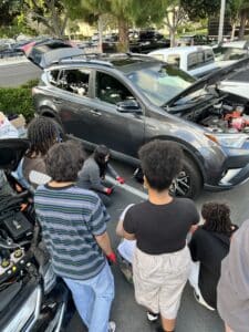 Students watch as one of their peers works on an auto in a parking lot.