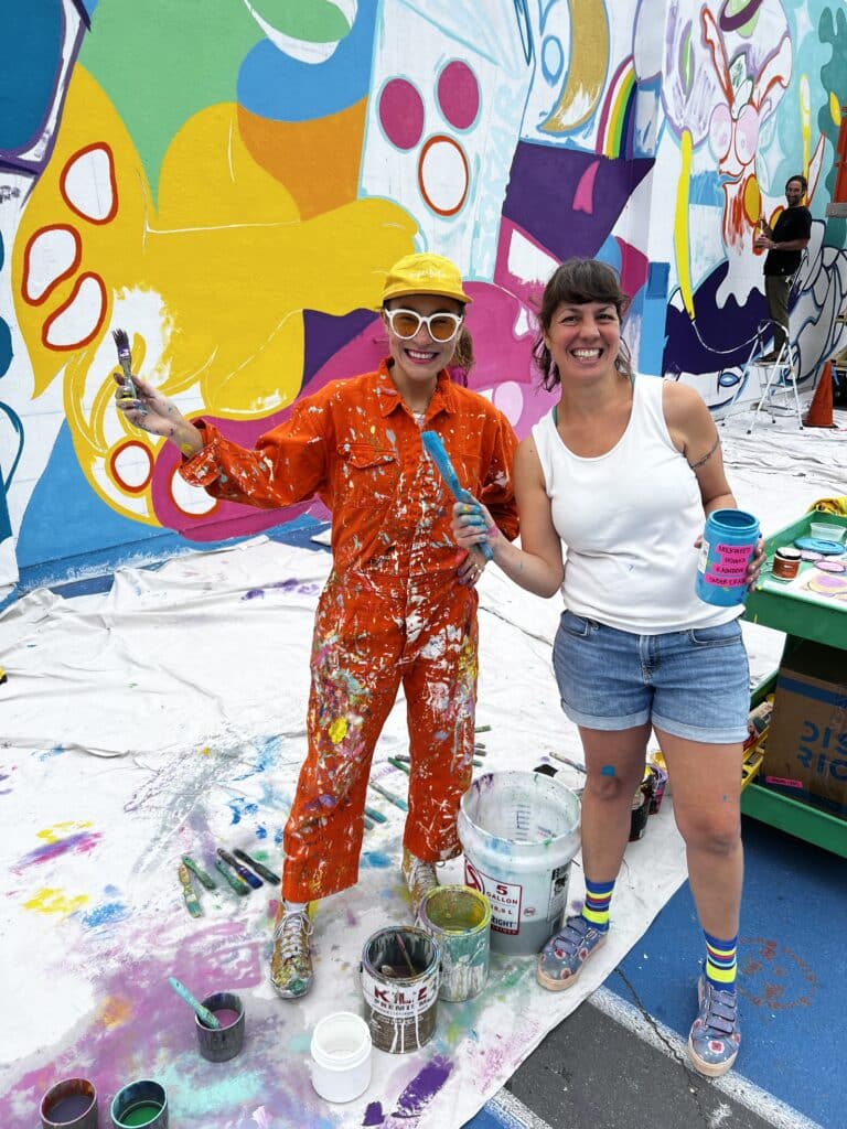 Visual artist Scarlett Baily poses in bright painter's outfit with a parent volunteer.