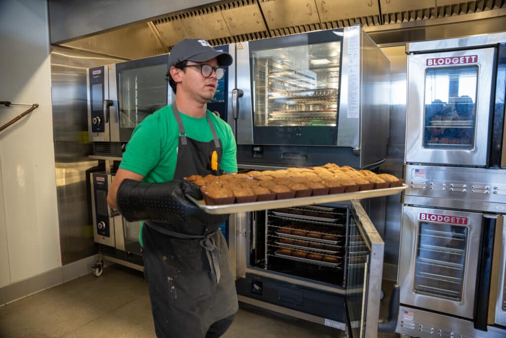 Food services staff carries a tray of muffins.